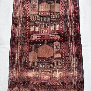 May include: A rectangular, hand-woven rug with a detailed, geometric design. The rug features a central pattern of stylized architectural elements in shades of brown, red, and beige, framed by a border with repeating patterns. The rug's dimensions are not visible.
