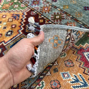 May include: A hand-woven area rug featuring a vibrant geometric pattern in shades of red, orange, blue, and green. The underside shows a light gray woven backing with white fringe tassels along the edge, revealing the construction of the textile.