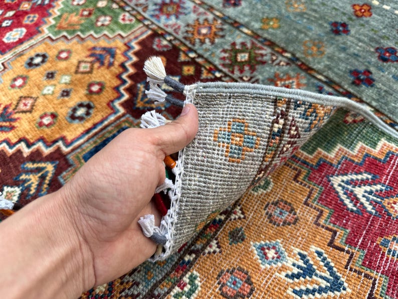 May include: A hand-woven area rug featuring a vibrant geometric pattern in shades of red, orange, blue, and green. The underside shows a light gray woven backing with white fringe tassels along the edge, revealing the construction of the textile.