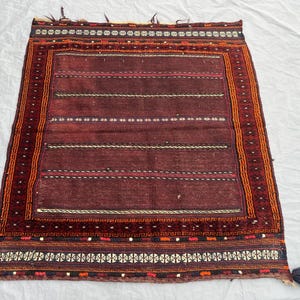 May include: A rectangular, hand-woven rug with a deep burgundy field and intricate borders. The rug features horizontal stripes in varying shades of red, white, and black. The borders are detailed with geometric patterns in orange, black, and white.