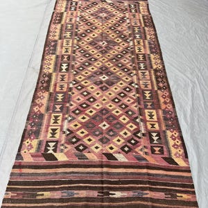 Puede incluir: Una alfombra rectangular tejida a mano con un patrón geométrico. La alfombra presenta un diseño central de diamantes en tonos marrones, rosas y amarillos, enmarcado por bordes con motivos geométricos y florales. La alfombra tiene un borde con flecos.