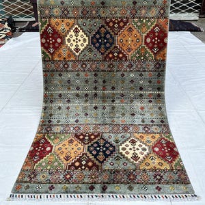 May include: A rectangular area rug featuring a central field of sage green with small multicolored floral motifs. The top and bottom borders display large geometric diamond patterns in shades of red, navy blue, orange, and green with white fringe.