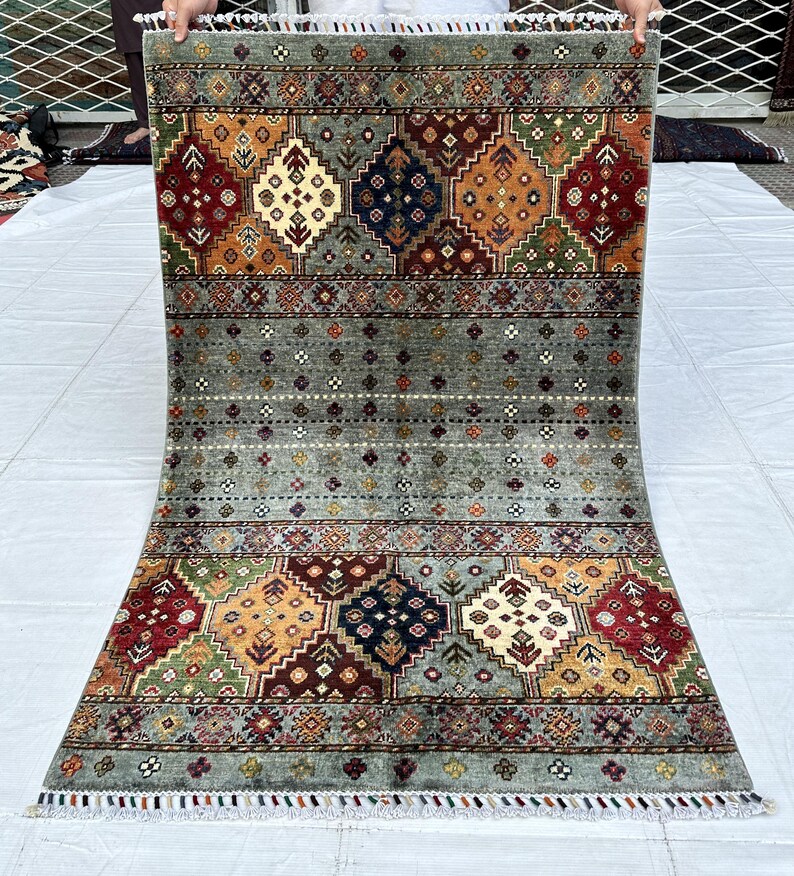 May include: A rectangular area rug featuring a central field of sage green with small multicolored floral motifs. The top and bottom borders display large geometric diamond patterns in shades of red, navy blue, orange, and green with white fringe.