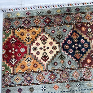 May include: A rectangular area rug featuring a repeating geometric pattern of diamonds in shades of red, gold, navy blue, and cream against a light blue background. The rug is finished with a short white fringe along the top edge.
