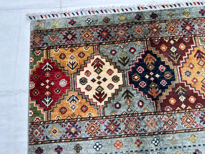 May include: A rectangular area rug featuring a repeating geometric pattern of diamonds in shades of red, gold, navy blue, and cream against a light blue background. The rug is finished with a short white fringe along the top edge.