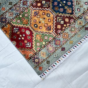 May include: A rectangular area rug featuring a geometric pattern of octagonal shapes in shades of red, orange, green, and blue. The rug has a light blue border with small floral motifs and a white fringe edge with multicolored thread accents.