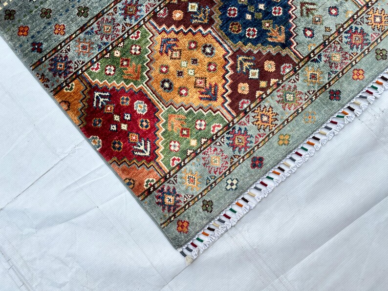 May include: A rectangular area rug featuring a geometric pattern of octagonal shapes in shades of red, orange, green, and blue. The rug has a light blue border with small floral motifs and a white fringe edge with multicolored thread accents.