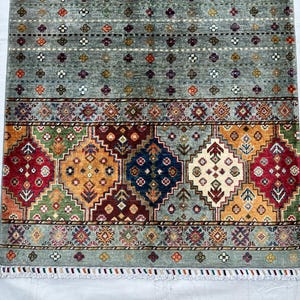 May include: A rectangular area rug featuring a gray field with small multicolored floral motifs. The lower section displays a wide border with large diamond-shaped patterns in red, blue, yellow, and green, finished with a short white fringe at the edge.