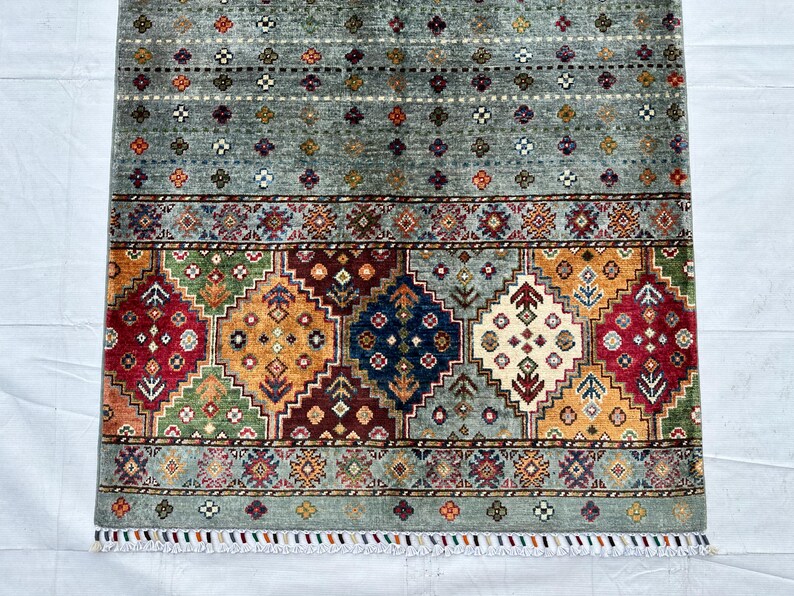 May include: A rectangular area rug featuring a gray field with small multicolored floral motifs. The lower section displays a wide border with large diamond-shaped patterns in red, blue, yellow, and green, finished with a short white fringe at the edge.