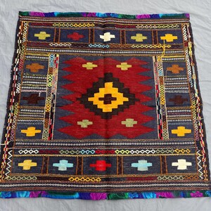 May include: Handmade square rug with a geometric design. The rug features a central red field with a black and yellow diamond pattern. The border is dark blue with colorful cross and geometric shapes.