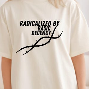 Può includere: T-shirt color crema con la scritta nera "RADICALIZED BY BASIC DECENCY" e una grafica nera. La maglietta ha maniche corte e una vestibilità ampia.