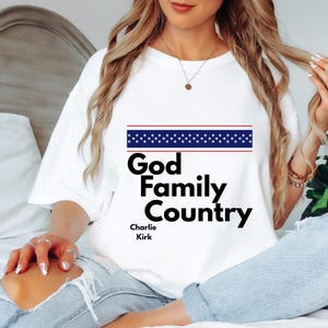 Puede incluir: Camiseta blanca con la frase "God Family Country" en negrita negra, con un diseño de rayas azules y rojas encima. El nombre "Charlie Kirk" está impreso debajo.