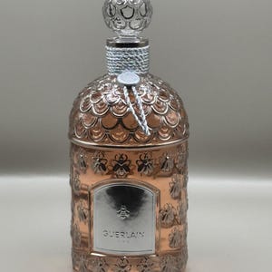 Könnte beinhalten: Kupferfarbener Parfümflakon mit einem dekorativen Bienenmotiv. Die Flasche hat einen transparenten, strukturierten Verschluss und ein silbernes Etikett mit dem Wort "GUERLAIN" und einem Bienenemblem. Die Flasche ist mit einer silbernen und weißen Kordel umwickelt.