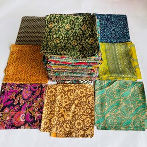 May include: A collection of colorful, patterned fabric squares in various designs. The fabrics include floral, paisley, and geometric patterns in shades of gold, green, blue, purple, and brown. The fabrics are stacked and arranged on a white surface.