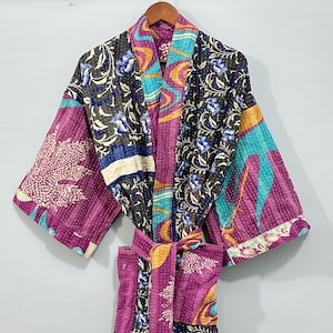 Peut inclure: Robe patchwork avec motifs floraux et géométriques dans les tons violets, turquoise et noirs. La robe a des manches longues et une ceinture assortie. Le tissu semble être matelassé, avec des coutures visibles sur tout le vêtement.