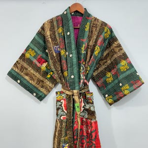 May include: A colorful, patterned robe with a belt, hanging on a wooden hanger. The robe features a mix of green, brown, red, and yellow fabrics with various designs. The interior lining is a bright pink. The robe is made of a soft, lightweight material.