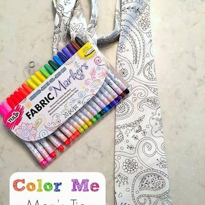 May include: A white tie with a black paisley design, next to a box of 20 fabric markers. The tie has the words "Color Me Men's Tie" on a white background. The markers are in various colors, including red, orange, yellow, green, blue, and purple.