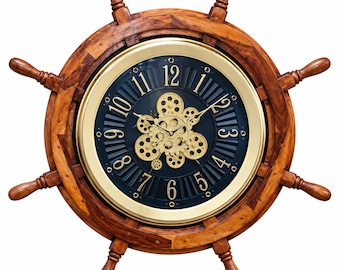 May include: A wall clock shaped like a wooden ship steering wheel with a polished gold-tone metal rim. The clock face features a dark blue background with gold-coloured Arabic numerals and a central cluster of exposed gold-tone gears.