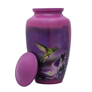 May include: A cylindrical metal cremation urn with a glossy magenta finish. The body features a printed graphic of a green hummingbird in flight alongside purple iris flowers. A matching circular magenta lid is positioned to the left side.
