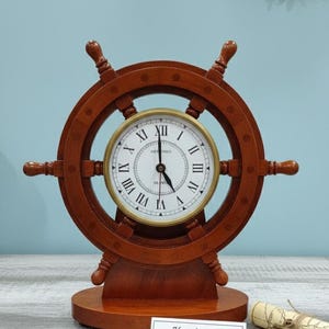 Könnte beinhalten: Eine braune Holzschiff-Steuerrad-Uhr mit weißem Zifferblatt und römischen Ziffern. Die Uhr hat ein maritimes Thema und ist mit "Handmade Coastal Decor" beschriftet.
