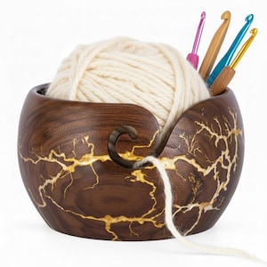 May include: A wooden yarn bowl with a dark brown finish and gold-colored crackle design. The bowl holds a ball of cream-colored yarn and several crochet hooks. The yarn exits the bowl through a curved slot.