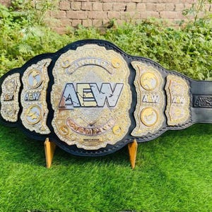 Könnte beinhalten: Ein Wrestling-Championgürtel mit einem schwarzen Lederriemen und goldenen und silbernen Akzenten. Die Mittelplatte zeigt "AEW" in Silber, umrahmt von Gold. Der Gürtel wird auf einer grünen Oberfläche präsentiert.