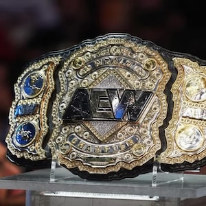 May include: A close-up of a wrestling championship belt. The belt features a large, ornate gold and silver center plate with the letters "AEW". The belt has a black leather strap and is displayed on a clear stand.