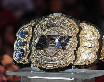 以下が含まれることがあります： プロレスのチャンピオンベルトのクローズアップ。ベルトは、「AEW」の文字が入った、大きく装飾された金と銀の中央プレートが特徴です。ベルトは黒革のストラップで、透明なスタンドに展示されています。