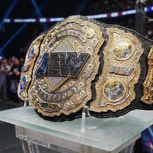 Może przedstawiać: Złoty i srebrny pas mistrzowski z widocznymi literami "AEW". Pas jest na przezroczystym stojaku, z czarnym skórzanym paskiem i skomplikowanymi detalami. Tło przedstawia arenę wrestlingu.