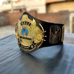 May include: A championship wrestling belt featuring a large, gold-colored metal center plate with an eagle emblem, a blue globe, and the text "WORLD HEAVYWEIGHT WRESTLING CHAMPION." The plate is attached to a black strap with additional gold side plates.