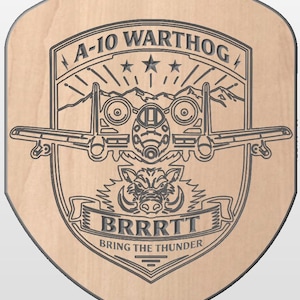 May include: A wooden plaque with a shield shape, featuring an engraved design of an A-10 Warthog aircraft, mountains, stars, and a wild boar. The text "A-10 WARTHOG", "BRRRTT", and "BRING THE THUNDER" are also engraved.