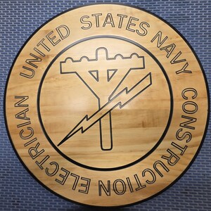 May include: A round wooden plaque with the words "UNITED STATES NAVY CONSTRUCTION ELECTRICIAN" in a circular pattern. The center features a black graphic of a utility pole and lightning bolt.