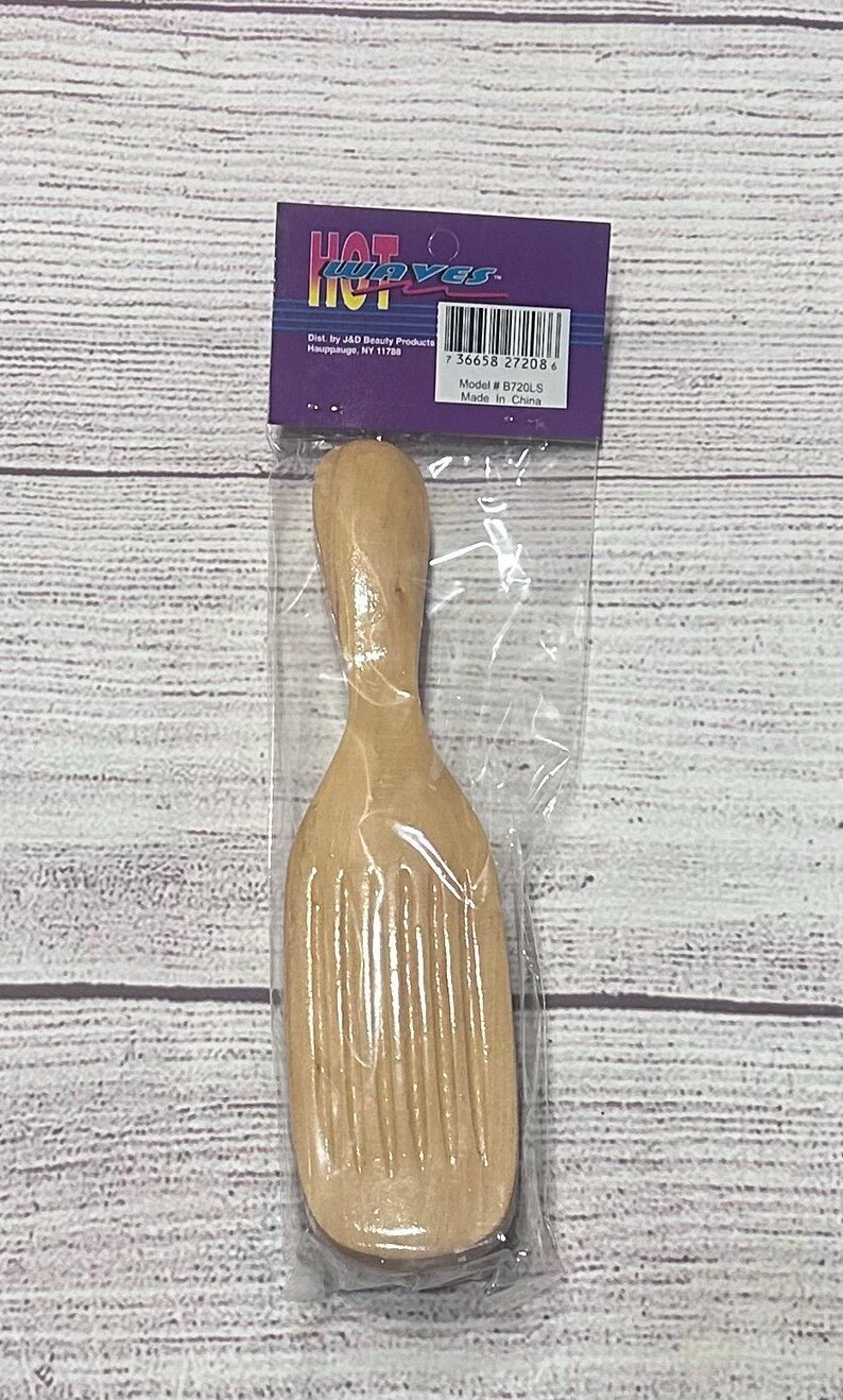 Puede incluir: Una herramienta de peinado de madera con un mango y una cabeza ancha y acanalada, empaquetada en una bolsa de pl&aacute;stico transparente. El embalaje incluye el texto "HOT WAVES" e informaci&oacute;n del producto. La herramienta est&aacute; fabricada en China.