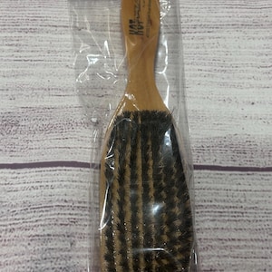 Puede incluir: Un cepillo de pelo de madera con cerdas oscuras, empaquetado en pl&aacute;stico transparente. El cepillo tiene un mango curvo y una cabeza rectangular. El embalaje tiene las palabras "HOT WAVES" en morado y amarillo.