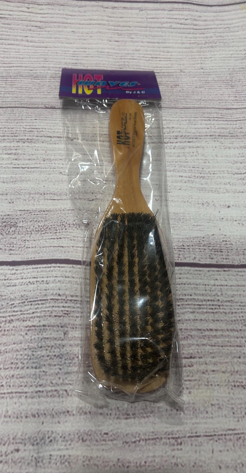 Puede incluir: Un cepillo de pelo de madera con cerdas oscuras, empaquetado en pl&aacute;stico transparente. El cepillo tiene un mango curvo y una cabeza rectangular. El embalaje tiene las palabras "HOT WAVES" en morado y amarillo.