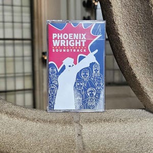 May include: A cassette tape case featuring the Phoenix Wright Ace Attorney Soundtrack. The cover art displays a white silhouette of a character pointing, set against a pink and blue background with white line art illustrations of various characters.