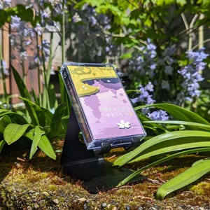May include: A cassette tape with the words "SPRING REIGN" and a floral design is displayed on a black metal stand. The tape's cover art features a landscape with purple, yellow, and green hues. The scene is set outdoors with green foliage and blue flowers in the background.