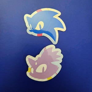 Puede incluir: Dos pegatinas con cabezas de personajes de dibujos animados. La pegatina superior es azul con una cara blanca y ojos amarillos. La pegatina inferior es morada con una cara blanca y ojos amarillos. Ambas pegatinas tienen un borde colorido sobre fondo azul.