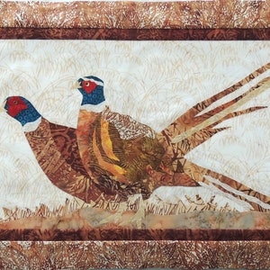 May include: Two brown and white pheasants with red and blue markings on their heads are perched on a brown and white background. The pheasants are made of fabric and are part of a quilt.