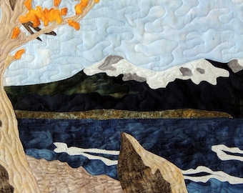 Puede incluir: Un tapiz de pared acolchado que presenta un paisaje estilizado con un árbol con hojas naranjas, un lago azul y una costa rocosa. El cielo es azul claro con nubes blancas.