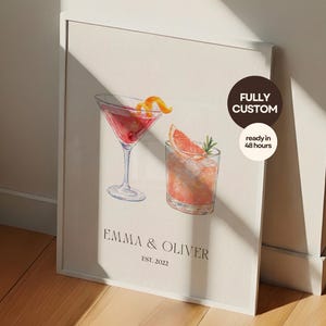 Puede incluir: Una impresión enmarcada con ilustraciones de acuarela de dos cócteles: un martini con una cáscara de naranja y una bebida con una rodaja de pomelo. La impresión incluye los nombres "Emma & Oliver" y "EST. 2022". Una pegatina circular dice "FULLY CUSTOM" y "ready in 48 hours".