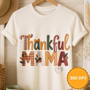 Könnte beinhalten: Ein weißes T-Shirt mit dem Schriftzug "Thankful Mama" in bunten Buchstaben im Patchwork-Stil. Das Design enthält herbstliche Akzente wie Blätter und ein Herz. Das Shirt wird auf einem Holzbügel präsentiert.