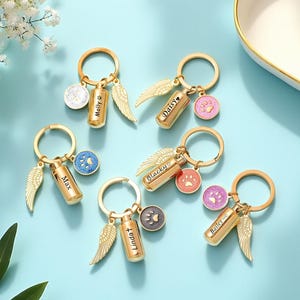 Puede incluir: Llaveros conmemorativos dorados con nombres grabados y dijes de alas de ángel. Cada llavero presenta un pequeño cilindro, un dije con una huella de pata y un dije circular. Los nombres Molly, Max, Daisy, Blacky, Linda y Bailey están grabados.
