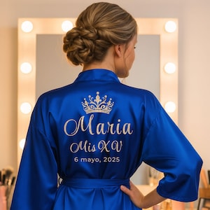 Könnte beinhalten: Königsblaue Satinrobe mit gesticktem Text und Kronenmuster. Der Text lautet "Maria Mis XV 6 mayo, 2025". Der Bademantel hat weite Ärmel und einen passenden Gürtel. Das Bild wird vor einem beleuchteten Schminkspiegel aufgenommen.