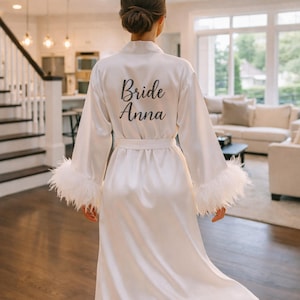 Pode incluir: Robe de cetim branco com acabamento de penas nas mangas. O robe tem um cinto a condizer e as palavras "Bride Anna" estão impressas nas costas em escrita preta. O robe vai até ao joelho.
