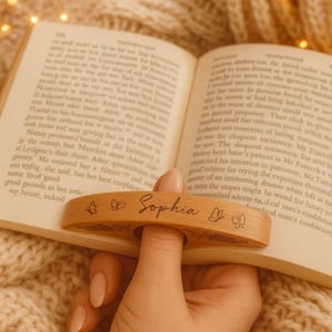 May include: A wooden book page holder with the name "Sophia" engraved, along with butterfly designs. The holder is designed to slip over a finger and keep a book open. The holder is shown resting on the pages of a book.