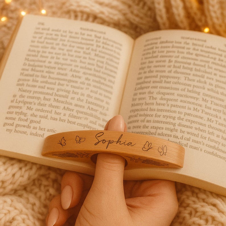 May include: A wooden book page holder with the name "Sophia" engraved, along with butterfly designs. The holder is designed to slip over a finger and keep a book open. The holder is shown resting on the pages of a book.