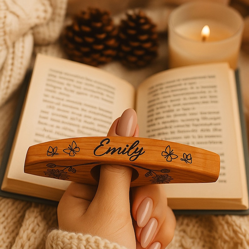 May include: A wooden book page holder with the name "Emily" engraved in black script. The holder features floral and butterfly designs. The holder is held over an open book, with a lit candle and pinecones in the background.