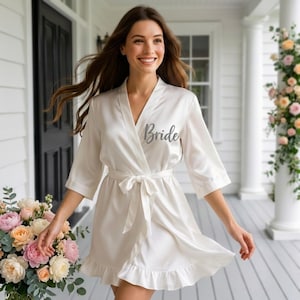 May include: Ivory satin robe with the word "Bride" in silver script. The robe has a wrap design with a tie belt and ruffled hem. The sleeves are three-quarter length. A bouquet of flowers is in the foreground.