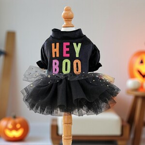 May include: A black pet dress featuring a mock neck and short sleeves. The bodice displays the text HEY BOO in colorful, glittery letters. The skirt consists of layered black tulle fabric adorned with small, shimmering gold star-shaped sequins.